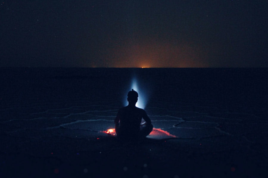 Siluetten av en person som sitter i mörkret framför en lägereld på en slätt och funderar över meningsfullhet